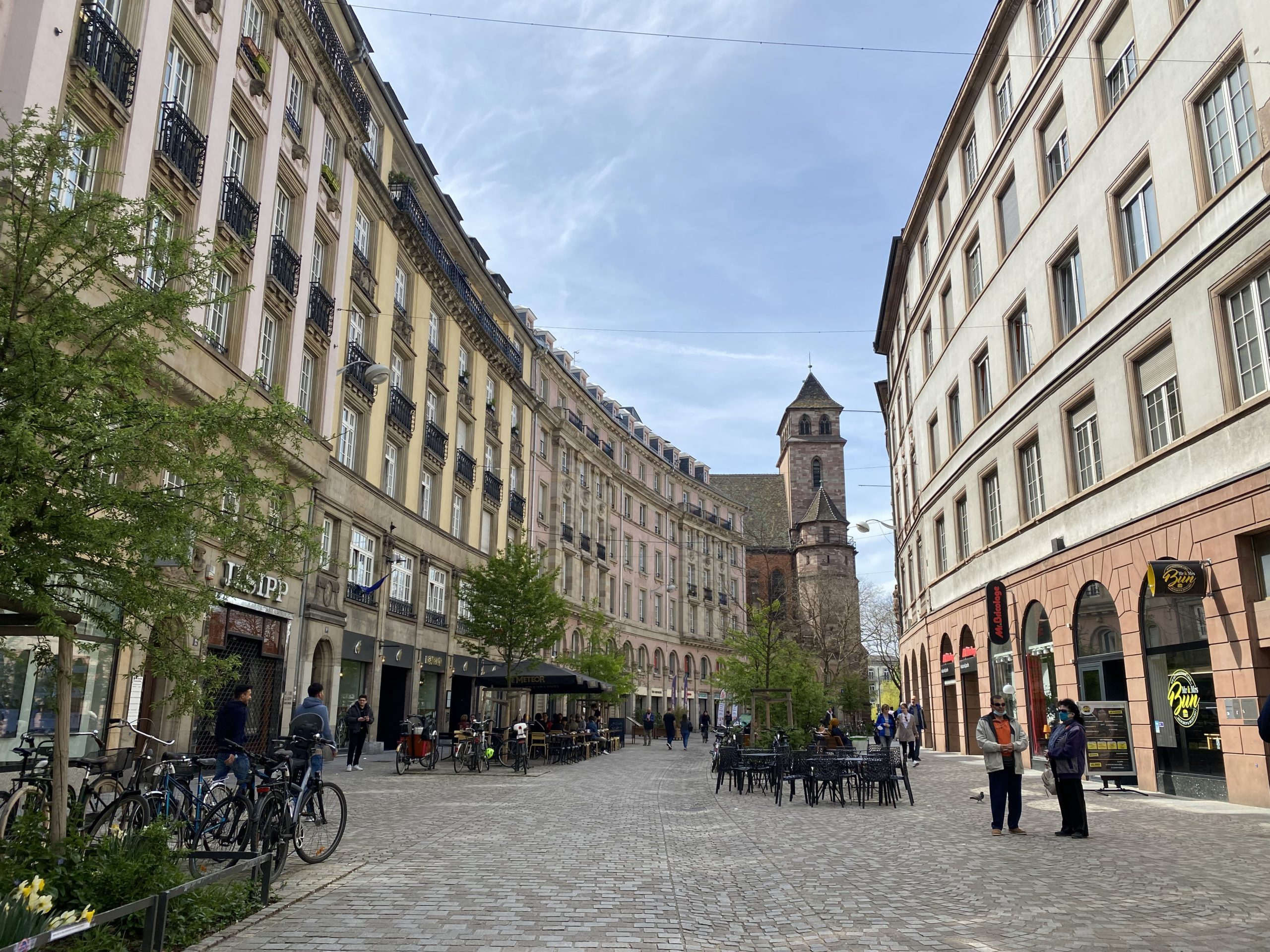 Centre-ville de Strasbourg comprenant une rue piétonne pavée, végétalisée et animée. Des personnes discutent. L'église est visible en arrière-plan