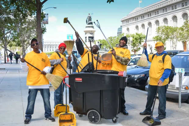 Cinq adultes, habillés d'un tshirt jaune floqué Downtown Streets Team et équipé d'outils de nettoyage, sourient à la caméra.