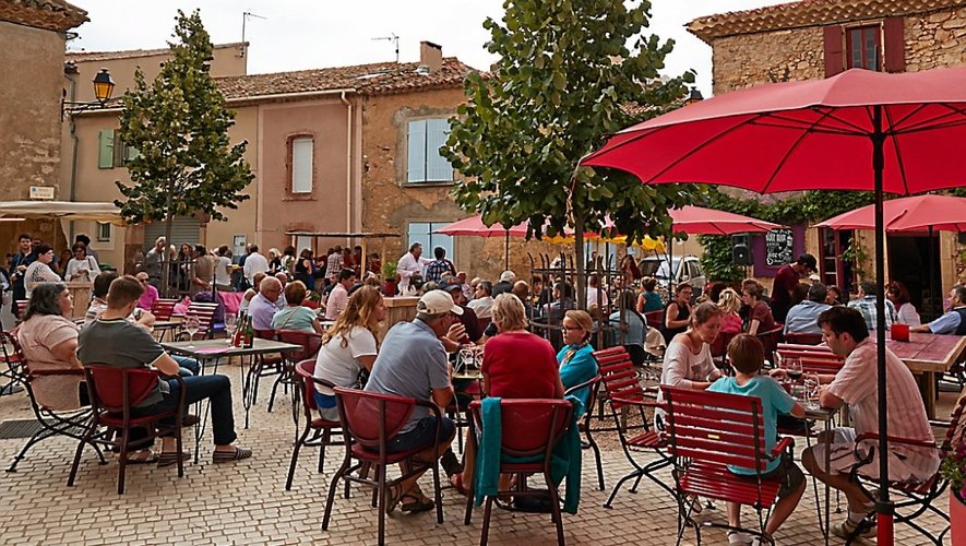 Place du centre-ville d'Assignan, de nombreuses personnes sont assises en terrasse d'un café.