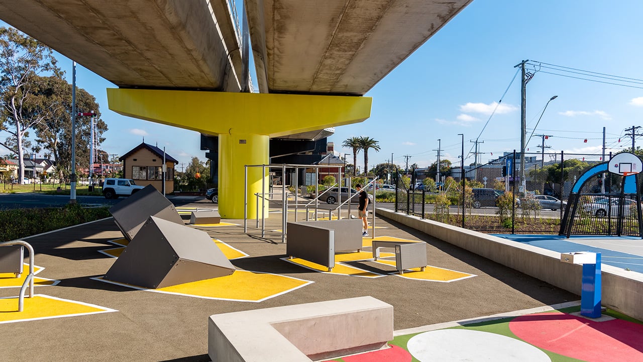 Sous la voie ferrée où les poteaux sont peints en jaune, il y a une aire de jeux avec du mobilier cubique et des marquages au sol