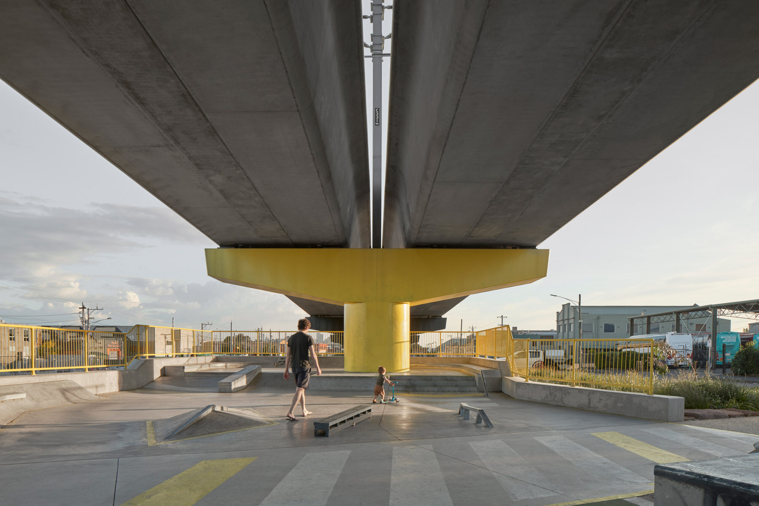 Sous la voie ferrée où les poteaux sont peints en jaune, présence d'un skatepark. Un enfant fait de la trottinette, accompagné d'un adulte