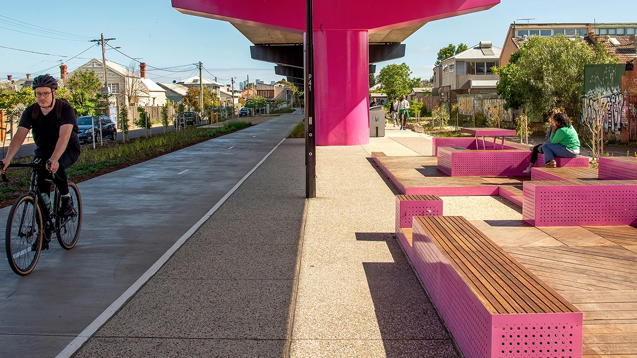 Sous la voie ferrée surélevée dont les poteaux sont peints en rose, il y a une piste cyclable à gauche et du mobilier rectangulaire en métal rose et bois à droite.