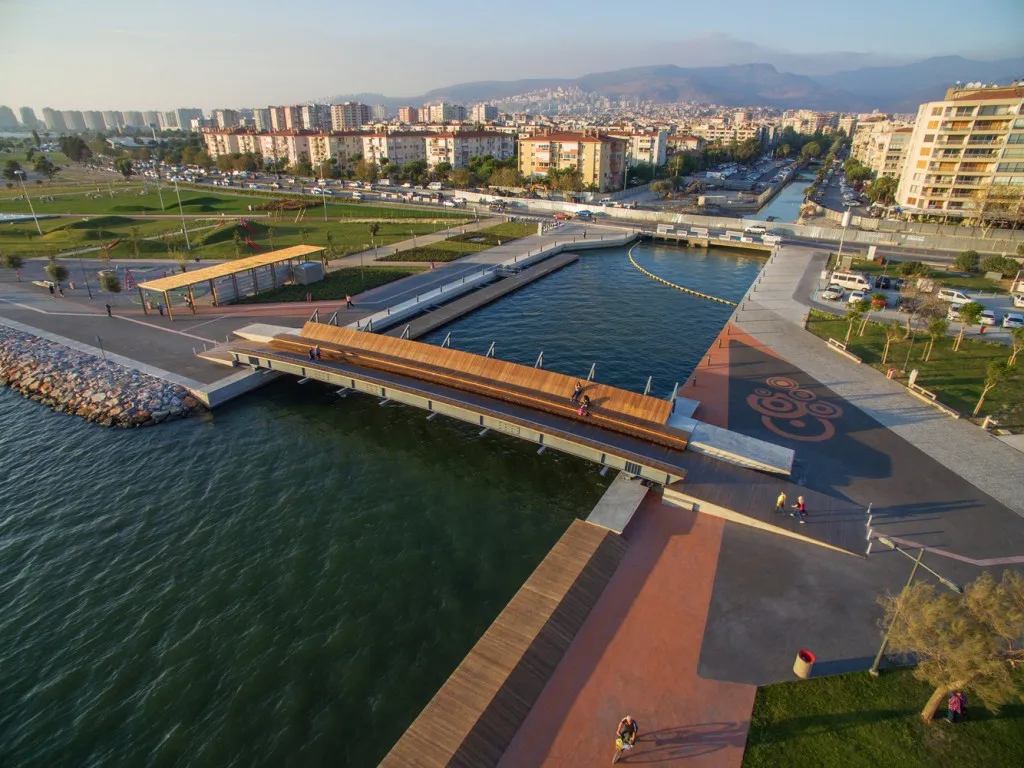 Passerelle en bois au premier plan, sur laquelle des personnes sont assises. En arrière-plan, la ville d'Izmir et des montagnes.