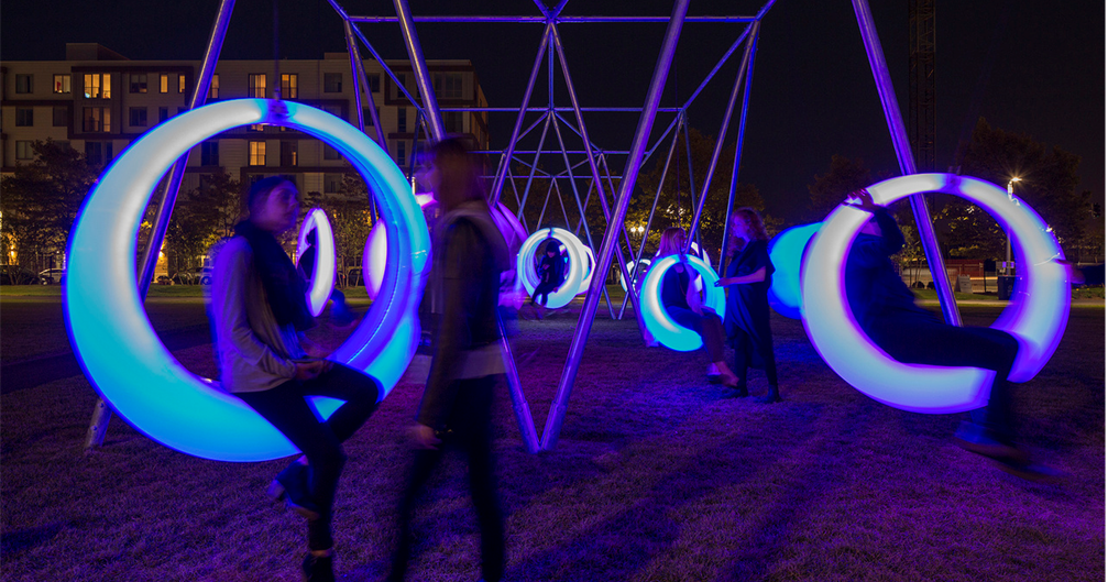 Structure métallique à laquelle sont accrochées des assises en forme d'anneaux qui sont allumés en bleu la nuit. Des personnes y sont assises.