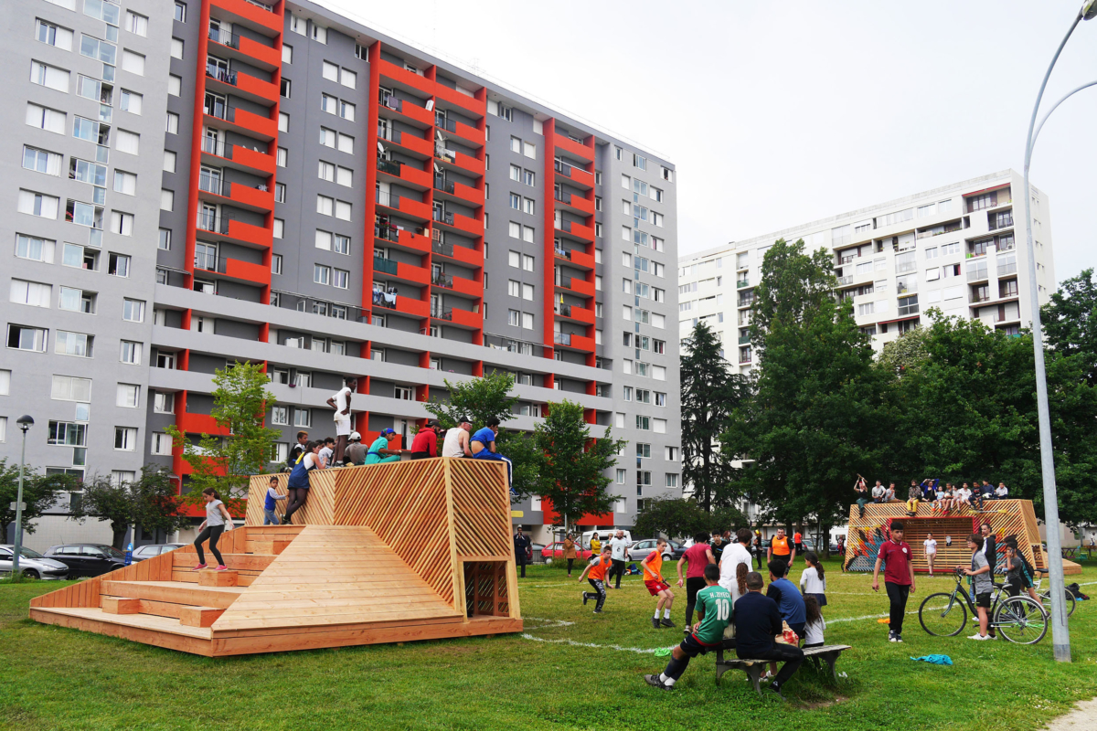 Terrain de foot matérialisé par du marquage au sol et des gradins en bois, au pieds d'immeubles